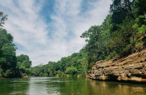 A serene river scene surrounded by lush green trees and rocky banks under a partly cloudy sky.
