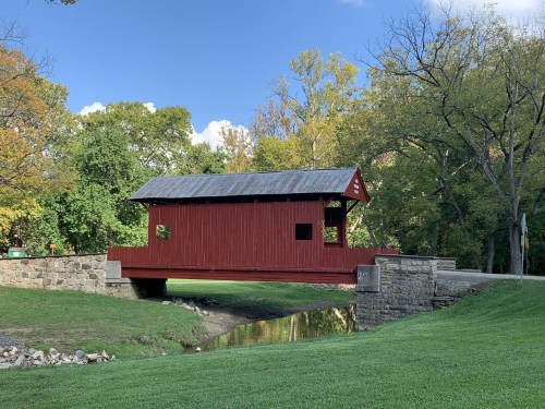 A red covered bridge spans a small stream, surrounded by trees and green grass under a clear blue sky.