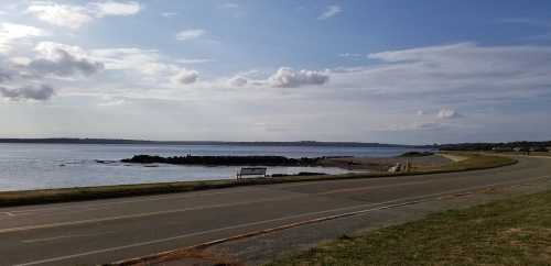 A serene coastal view with a calm sea, distant land, and a road lined with grass under a partly cloudy sky.