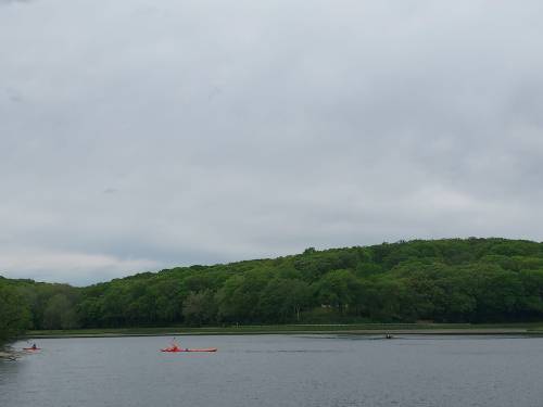 Two kayakers paddle on a calm lake surrounded by lush green hills under a cloudy sky.