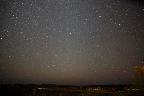 A starry night sky filled with countless stars over a dark landscape, with a faint horizon line visible.