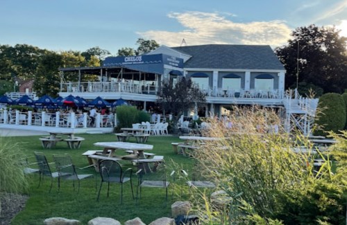 A waterfront restaurant with outdoor seating, surrounded by greenery and picnic tables, under a clear blue sky.