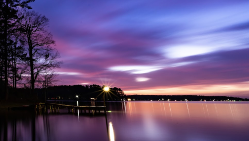 A serene lake at sunset, with colorful clouds reflecting on the water and a dock illuminated by soft lights.