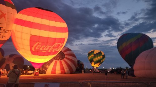 Colorful hot air balloons illuminated at dusk, with a cloudy sky and spectators in the foreground.
