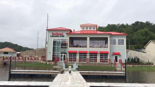 A modern lakeside restaurant with a red roof, large windows, and a dock in front, surrounded by greenery.