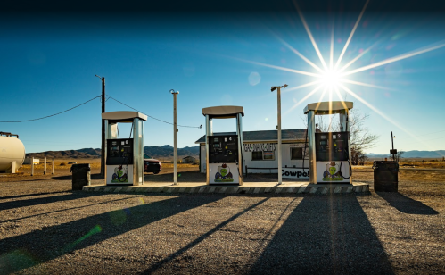 Abandoned gas station with three pumps, a small building, and mountains in the background under a bright sun.