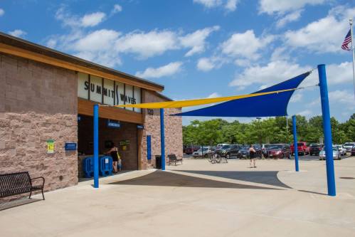 Entrance of Summit Waves water park with colorful shade sails, benches, and parked cars under a blue sky.