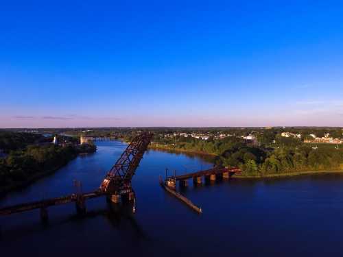 Aerial view of a river with a raised bridge, surrounded by greenery and a small town under a clear blue sky.