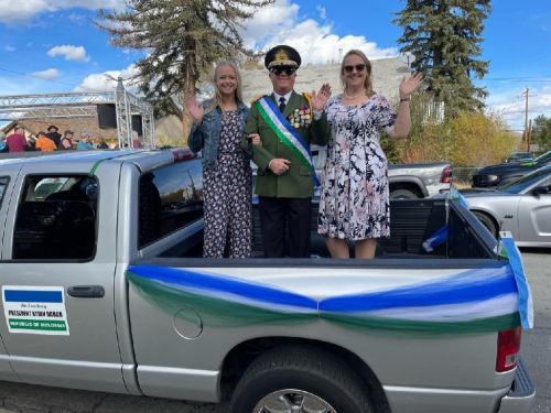 Three people wave from the back of a decorated pickup truck during a parade, with trees and blue sky in the background.