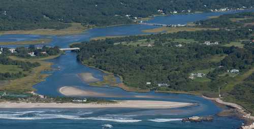 Aerial view of a winding river surrounded by lush greenery and sandy shores, with houses dotting the landscape.