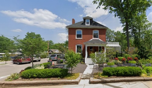 A red brick house with a porch, surrounded by greenery and colorful flower beds, on a sunny day.