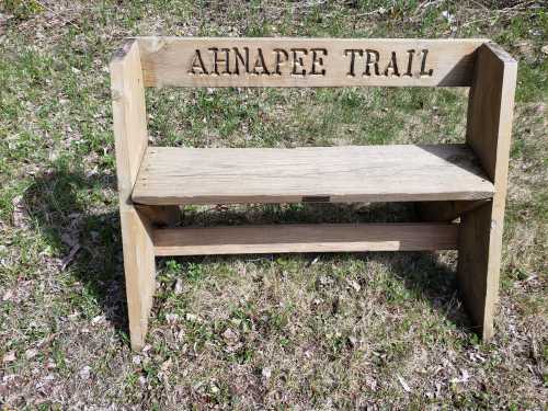 Wooden bench with "AHNAPEE TRAIL" carved on the back, set in a grassy area.