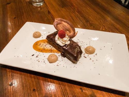 A chocolate dessert on a white plate, topped with whipped cream, a raspberry, and decorative elements, on a wooden table.