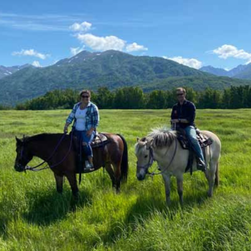 Two people on horseback in a green field with mountains in the background under a blue sky with fluffy clouds.