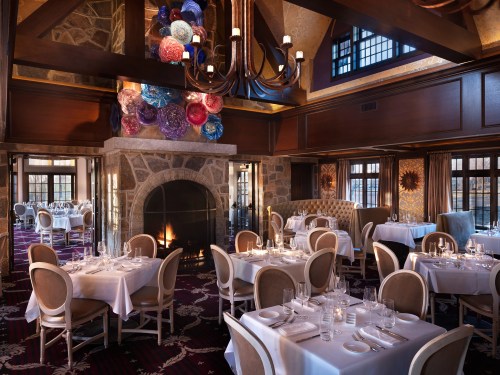 Elegant restaurant interior with tables set for dining, a stone fireplace, and colorful decor hanging from the ceiling.