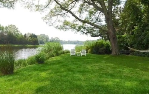 A serene riverside scene with green grass, a large tree, and two white chairs facing the water.
