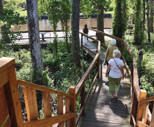 Two women walk down a wooden path surrounded by greenery, heading towards a river in a serene outdoor setting.