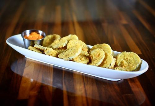 A white platter holds crispy fried pickles with a small cup of dipping sauce on a wooden table.