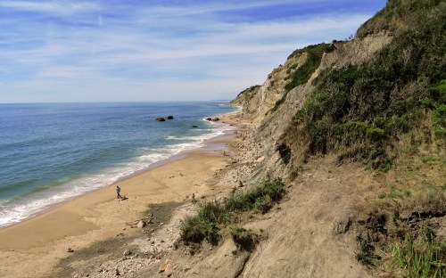 A serene beach scene with a person walking along the shore, cliffs in the background, and a clear blue sky.