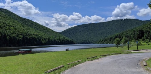 A serene lake surrounded by green mountains under a blue sky with fluffy clouds, featuring a grassy area and a road.