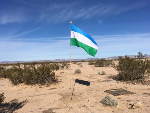 A blue and green flag stands on a pole in a desert landscape with sparse vegetation and distant mountains.