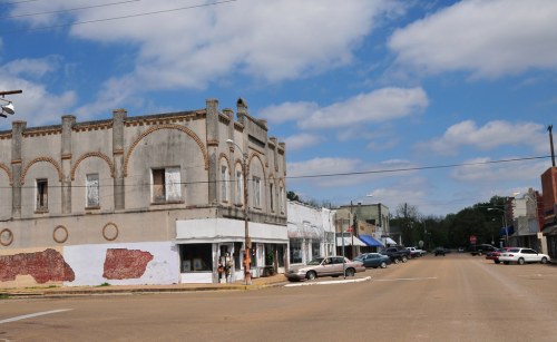 A small town street with a historic building, parked cars, and a blue sky with scattered clouds.