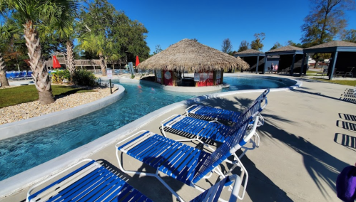 A tropical pool area with a thatched-roof bar, lounge chairs, and palm trees under a clear blue sky.