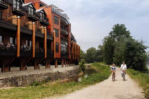 Two cyclists ride along a path beside a modern building with wooden accents, surrounded by greenery and a calm waterway.