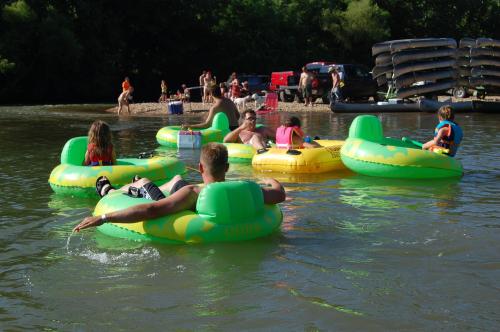 People relaxing on green inflatable rafts in a river, with others enjoying the beach and activities nearby.