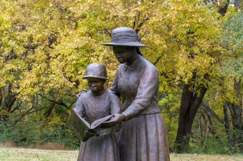A bronze statue of a woman and a child reading together, surrounded by autumn trees.