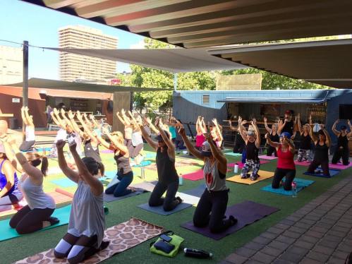 A group of people practicing yoga outdoors, kneeling with arms raised, on colorful mats under a shaded area.