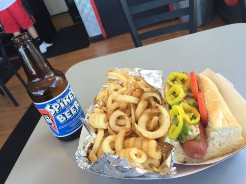A hot dog topped with yellow peppers and tomatoes, served with curly fries and a bottle of Spike Beer on a table.