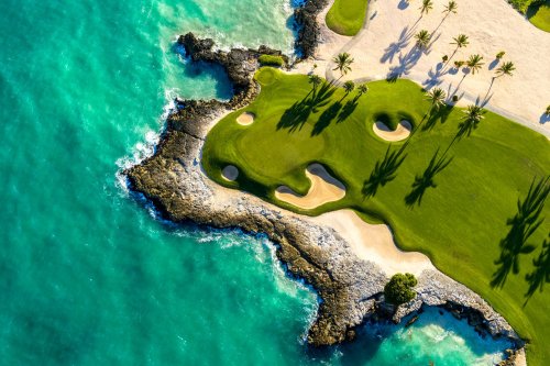 Aerial view of a golf course by the ocean, featuring sandy bunkers and palm trees casting shadows on the green.