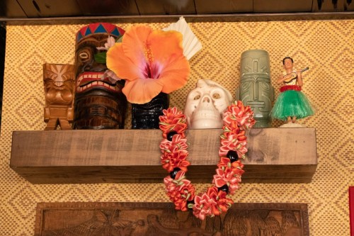 A wooden shelf displays colorful tiki mugs, a hibiscus flower, a skull, a hula dancer figurine, and a floral lei.