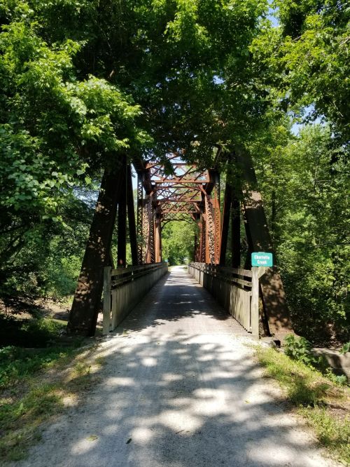 A rustic metal bridge surrounded by lush green trees, with a gravel path leading through it.