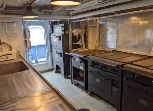 A ship's kitchen featuring stainless steel surfaces, multiple stoves, and a mixer, with a window in the background.