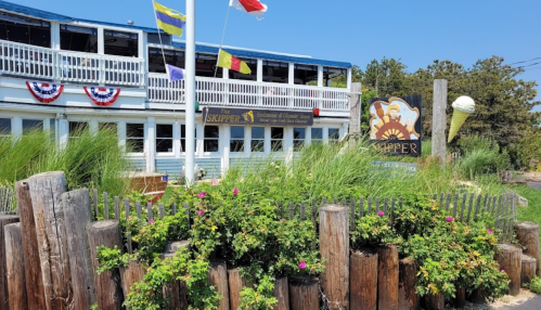 A seaside restaurant with colorful flags, greenery, and a sign reading "Skipper," featuring a sunny outdoor atmosphere.