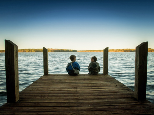 Two children sit on a wooden dock, gazing out at a calm lake surrounded by trees under a clear sky.