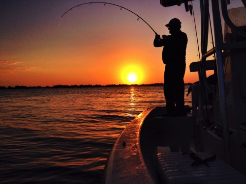 A silhouette of a person fishing on a boat at sunset, with vibrant orange and yellow hues reflecting on the water.