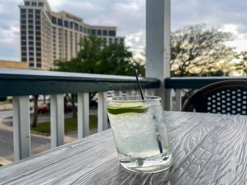 A refreshing cocktail with a lime wedge sits on a wooden table, with a hotel and trees in the background.