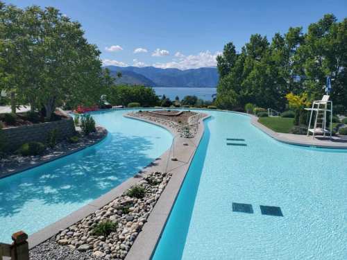 A scenic pool area with winding water features, surrounded by greenery and mountains under a clear blue sky.