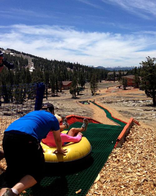 A person pushes a child on a yellow inflatable tube down a grassy slope, surrounded by trees and a clear blue sky.