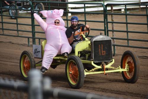 A man in a pig costume rides in a vintage car with another man, both smiling at a dirt track event.
