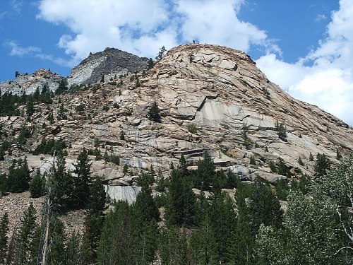 A rocky mountain slope with trees in the foreground and a blue sky with clouds above.