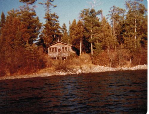 A rustic cabin nestled among trees on a rocky shoreline, viewed from a lake. Autumn colors surround the scene.