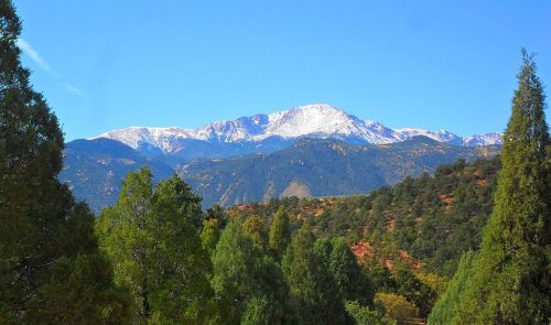 A scenic view of snow-capped mountains rising above lush green trees under a clear blue sky.