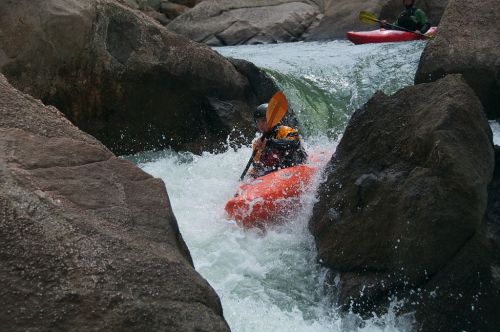 A kayaker navigates through rocky rapids, splashing water around them, with another kayaker in the background.