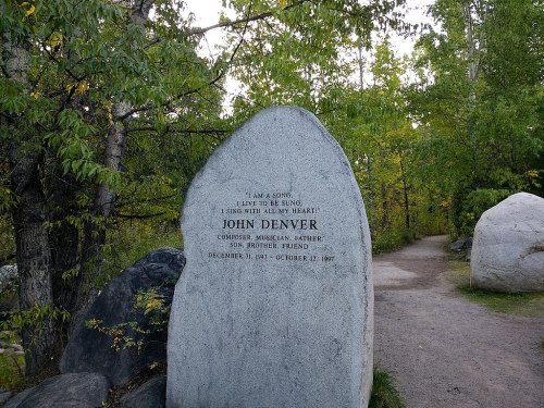 A stone memorial for John Denver, featuring an inscription and surrounded by trees along a pathway.