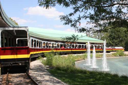 A colorful train curves around a pond with fountains, surrounded by greenery and a blue sky.