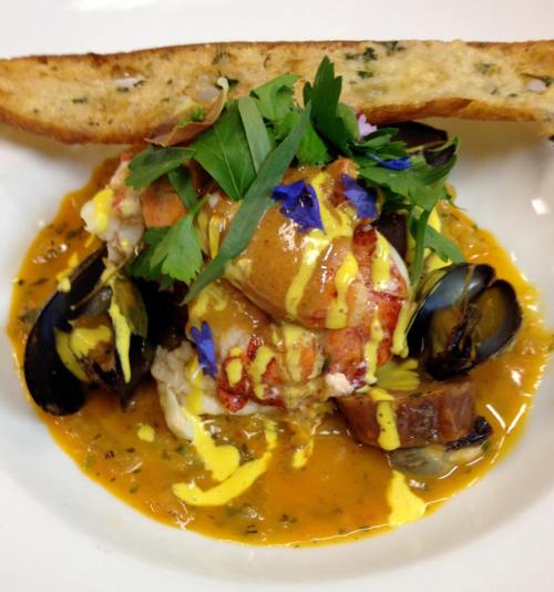 A gourmet seafood dish featuring lobster, mussels, and herbs, served with a slice of garlic bread.